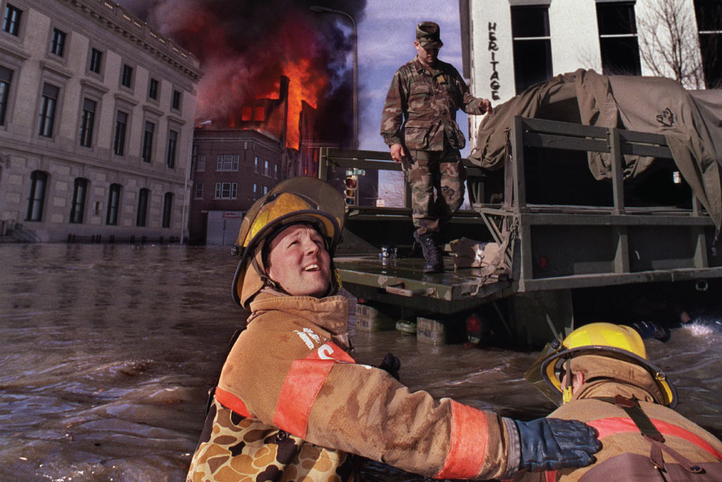 firefighters stand in flooded water while watching flames on buildings behind them